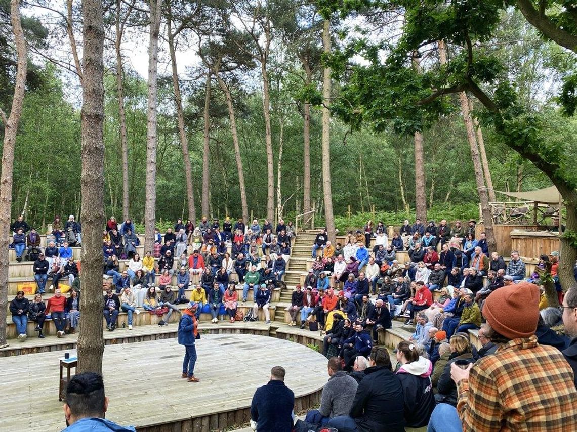 People sitting in an outdoor theatre in the forest watching a man on stage.