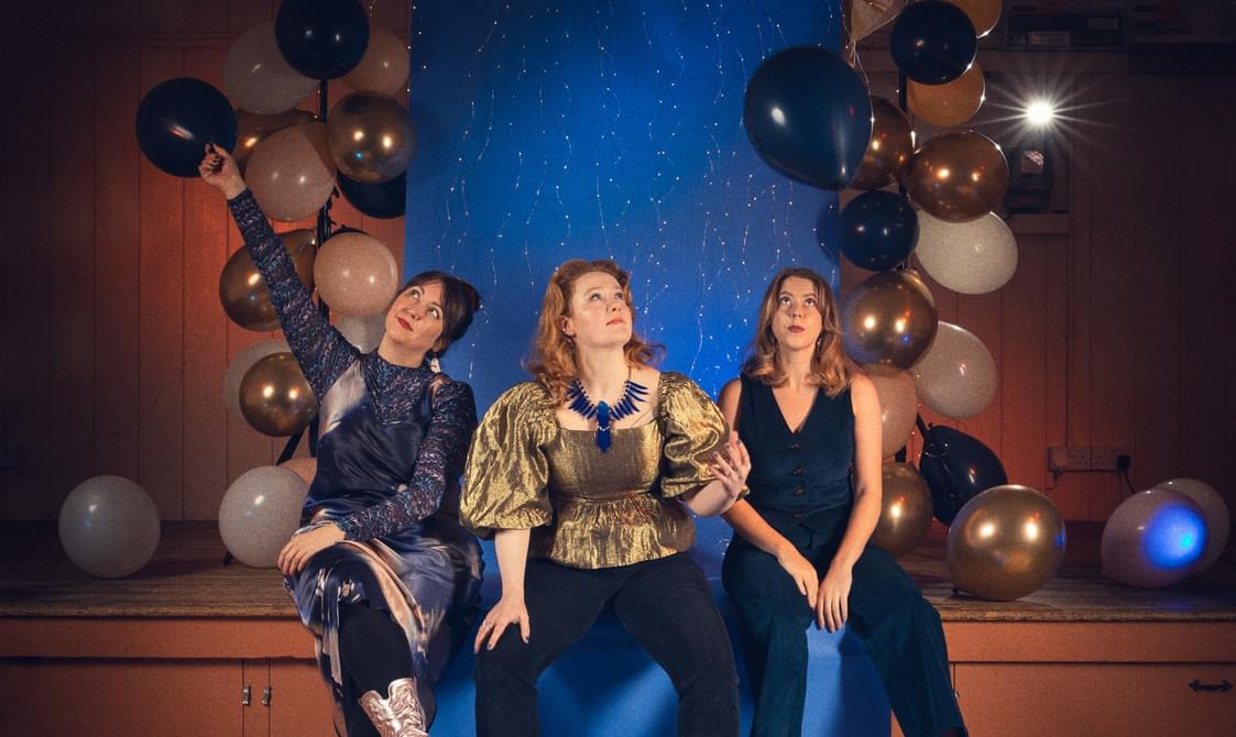 Three women wearing sparkly clothes sitting on a stage surrounded by balloons.