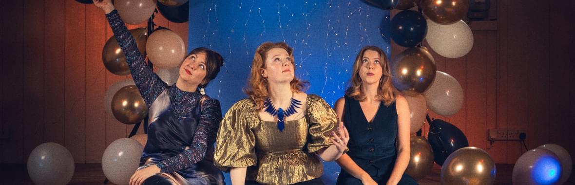 Three women wearing sparkly clothes sitting on a stage surrounded by balloons.
