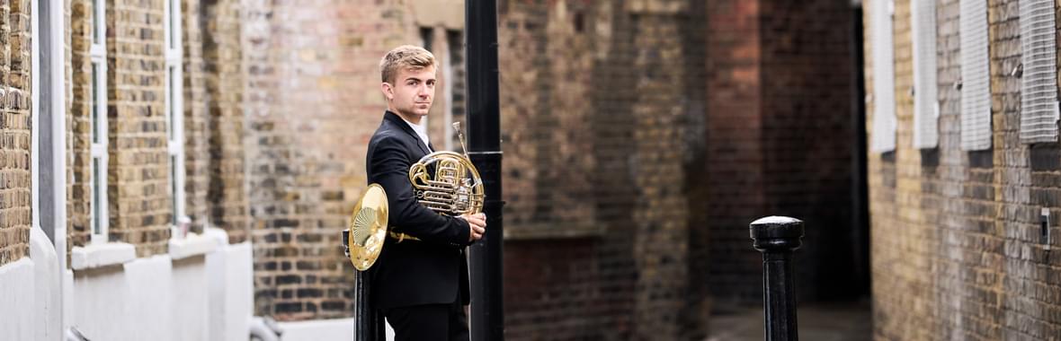 Man with blond hair wearing a dark suit and holding a French horn, standing in a quaint city lane.