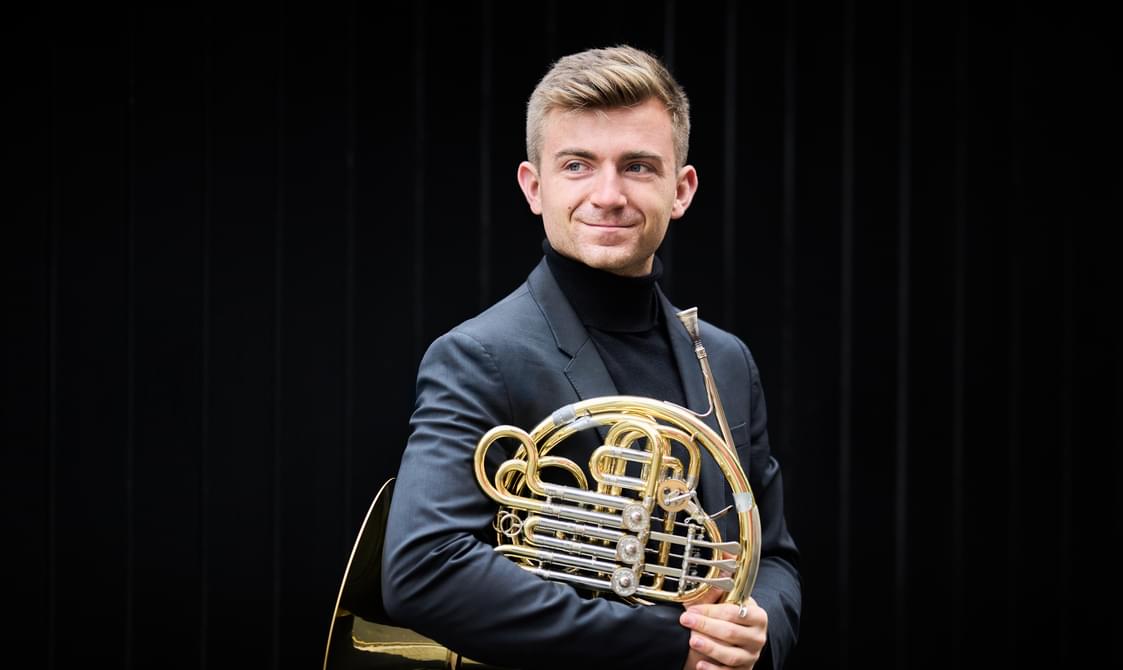 Man with blond hair wearing a black jacket and holding a French horn, against a dark background.