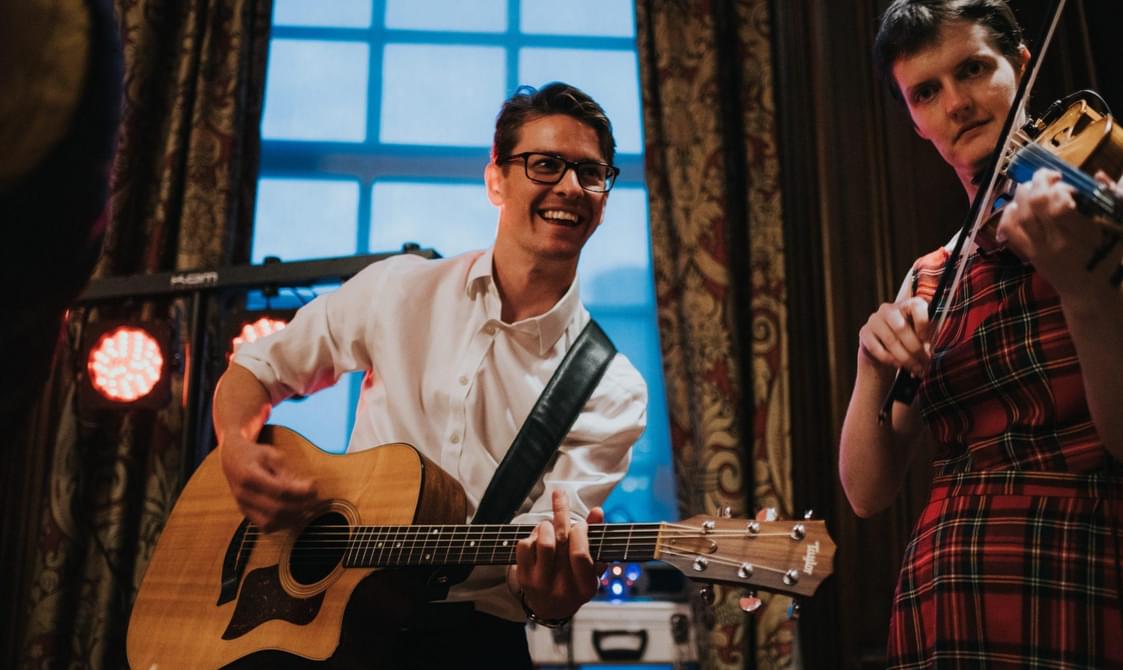 Man smiling and playing the guitar beside woman playing a violin.