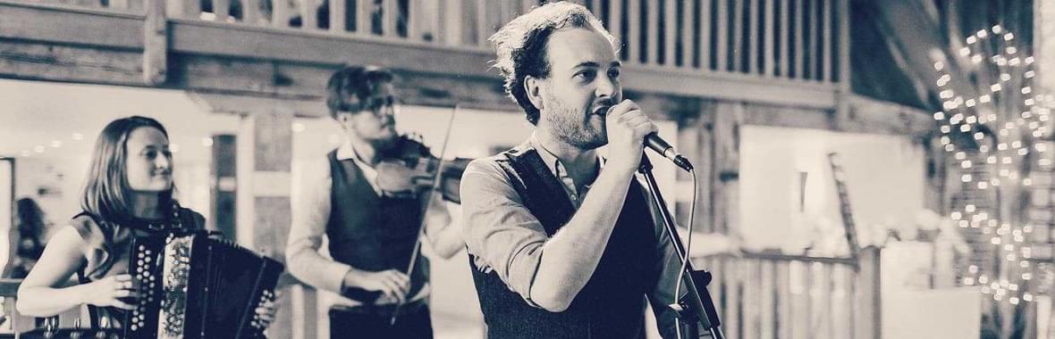 Black and white view of a man singing, man playing violin and woman playing the accordion in a barn with fairy lights.