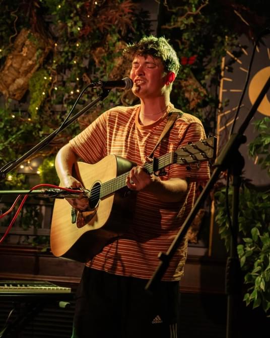 Young man with short hair wearing a tshirt, singing and playing guitar.