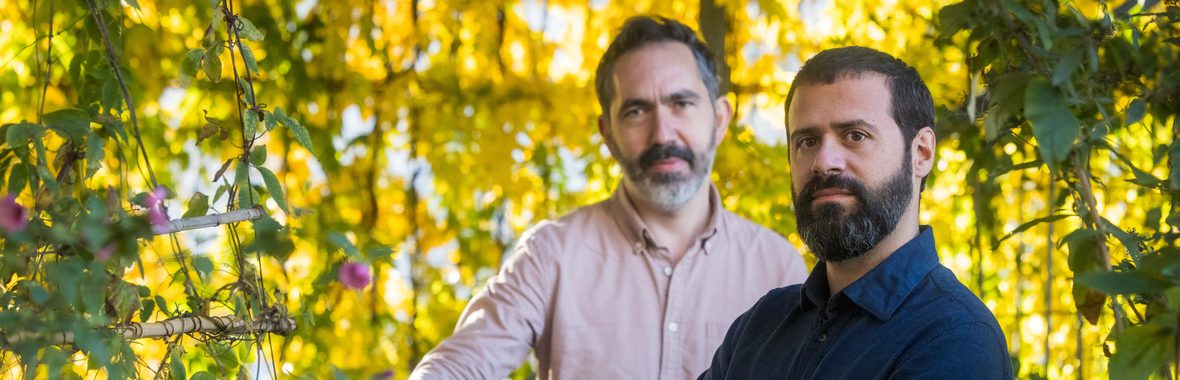 Two men with beards standing amidst greenery and flowers.
