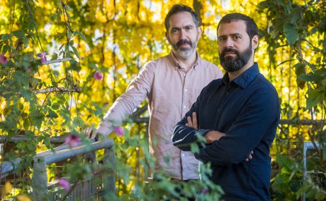 Two men with beards standing amidst greenery and flowers.