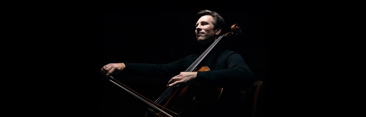 Man with short hair playing the cello against a black background.