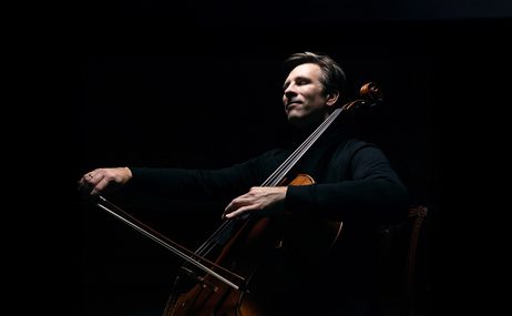 Man with short hair playing the cello against a black background.