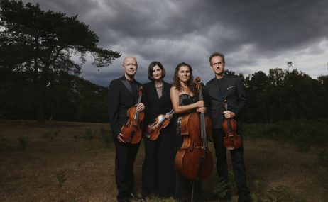 Two men and two women wearing formal dress, standing outside in a dark forest, holding their string instruments.