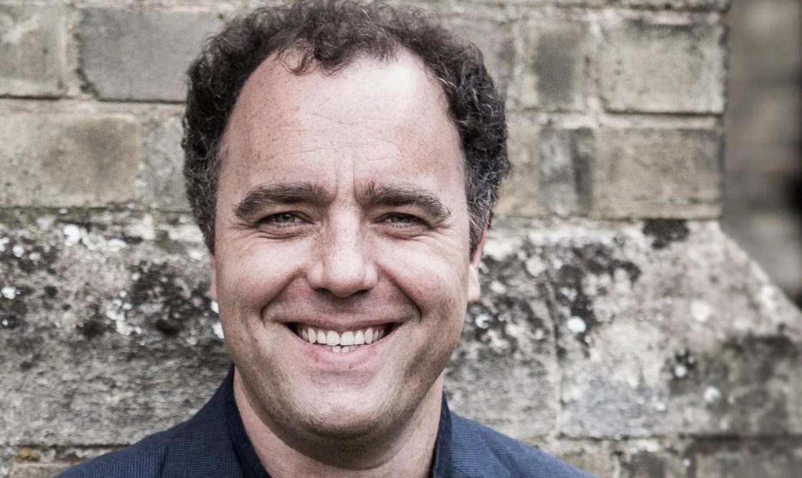Man with short black curly hair standing against a brick wall and smiling.