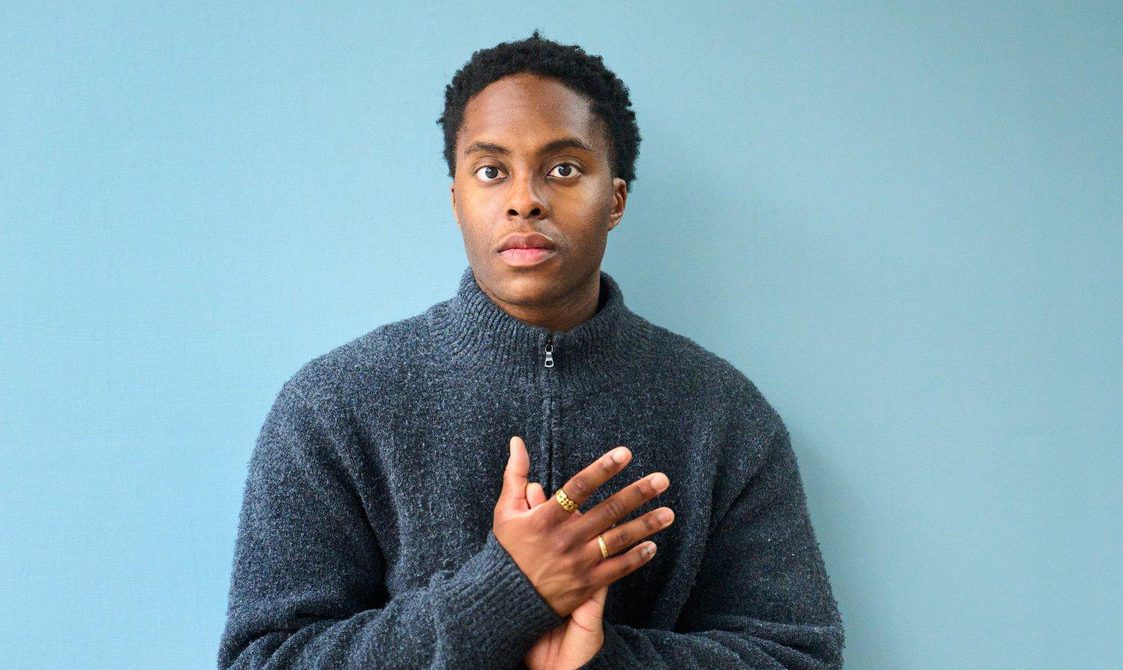Young man with short black hair wearing a blue sweater standing against a light blue background.