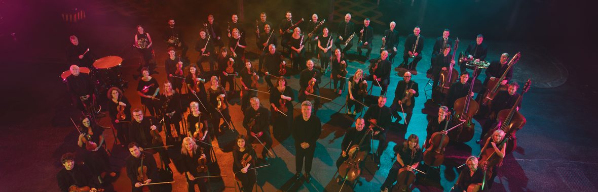 Bird's eye view of BBC Scottish Symphony Orchestra with Ryan Wigglesworth, standing holding their instruments under multicoloured lighting.