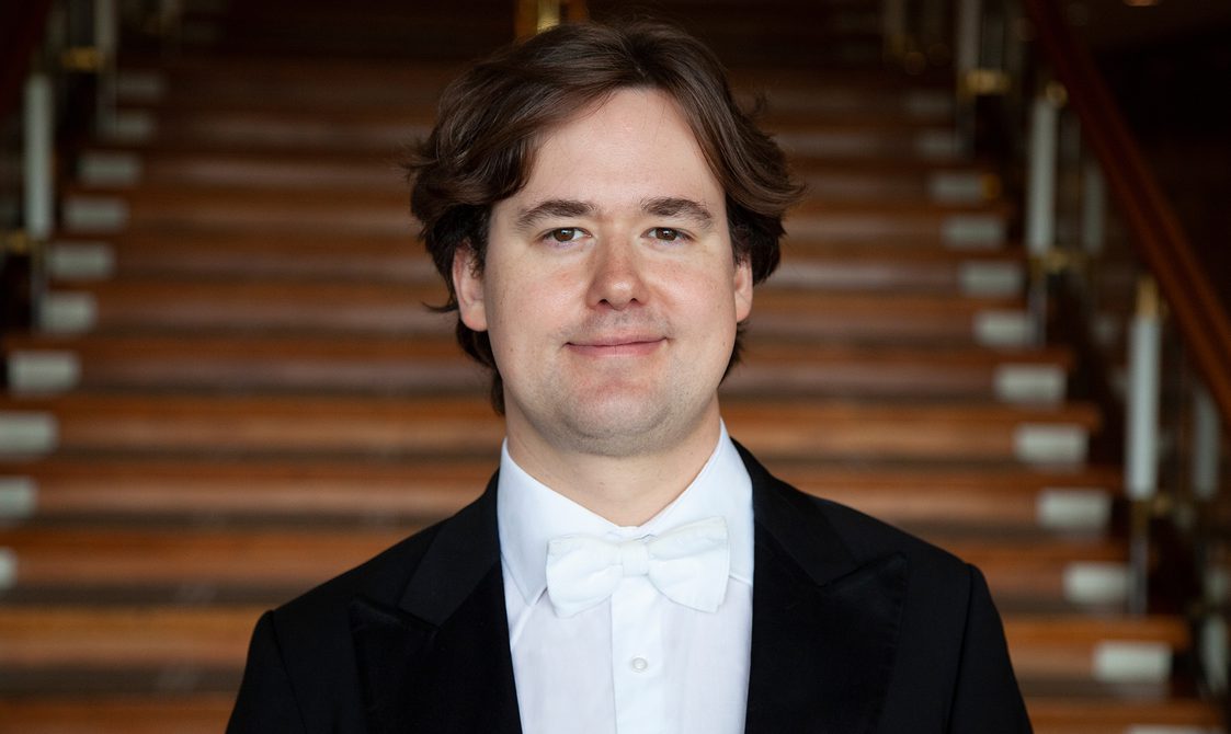 Man with short brown hair wearing a black jacket, white shirt and white bow tie.