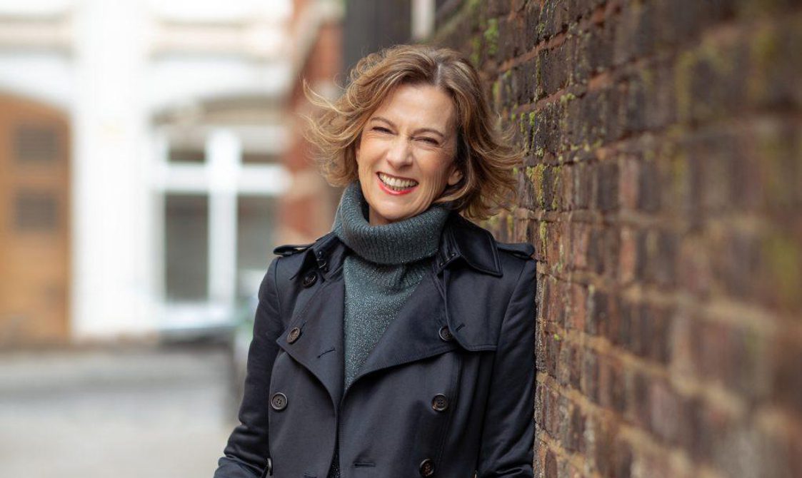 Woman with light brown bobbed hair, wearing a blue polo neck and raincoat, leaning against a brick wall and smiling.