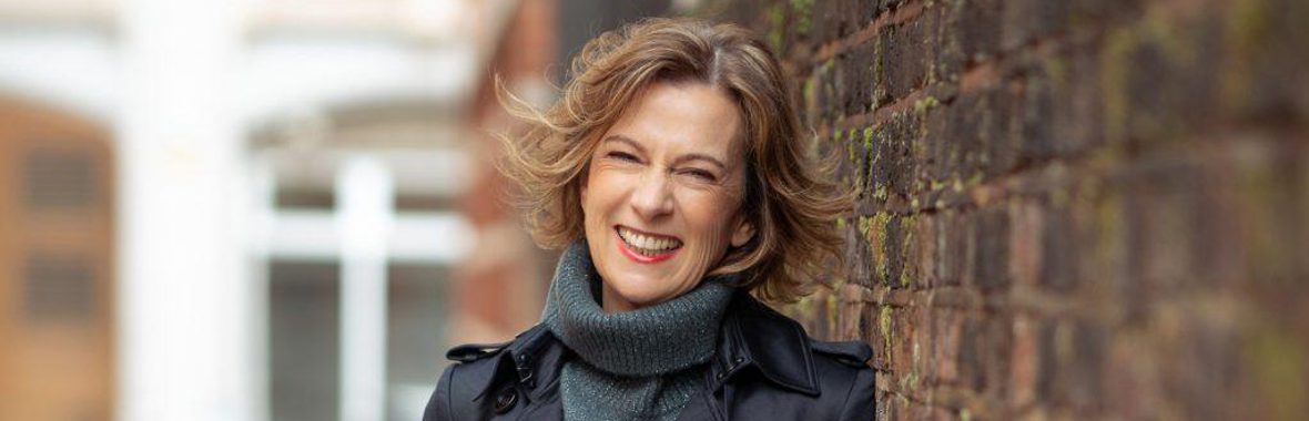Woman with light brown bobbed hair, wearing a blue polo neck and raincoat, leaning against a brick wall and smiling.