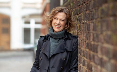 Woman with light brown bobbed hair, wearing a blue polo neck and raincoat, leaning against a brick wall and smiling.