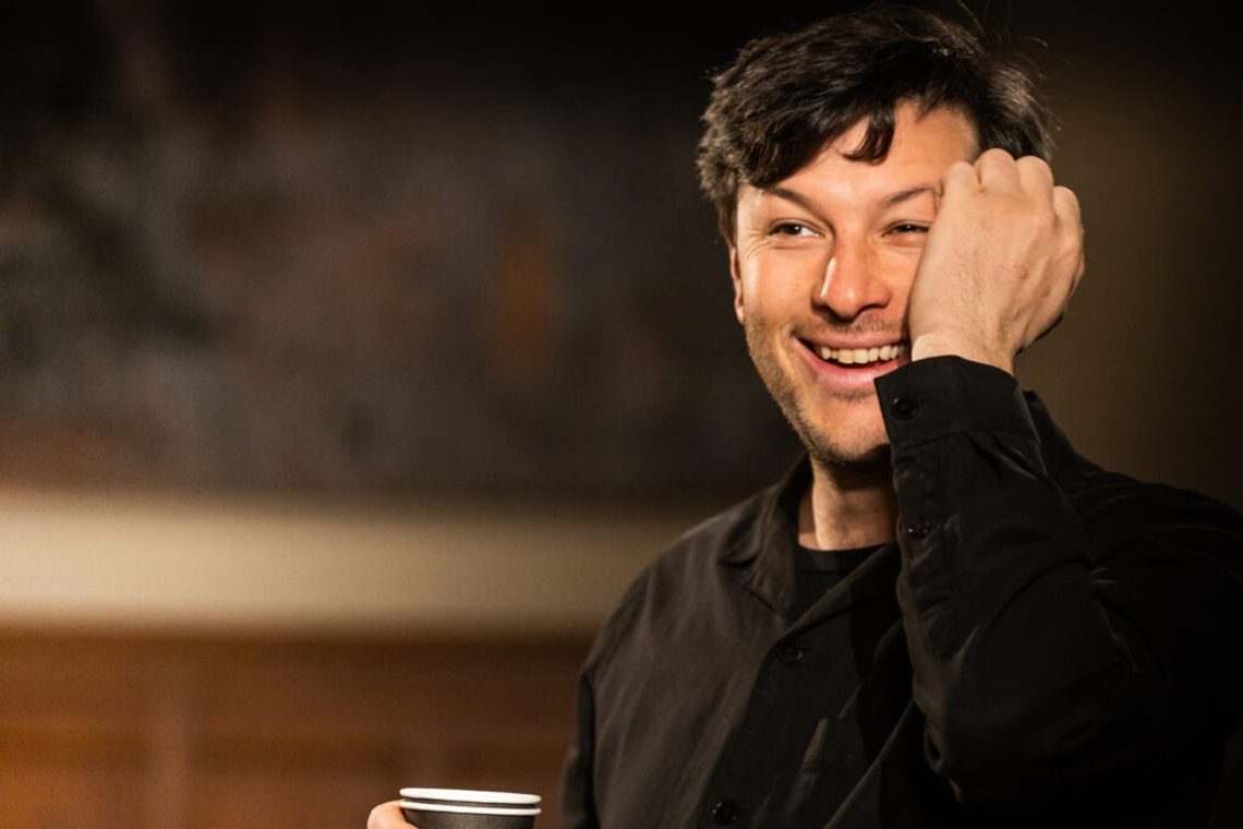Man with short dark hair wearing a dark top and holding his hand to his face and smiling.