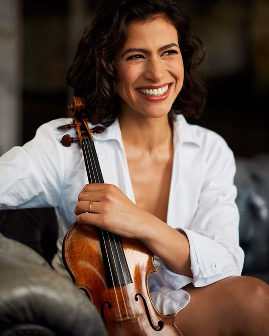 Woman with dark hair wearing a white shirt, holding a violin and smiling.