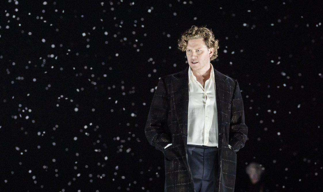 Man with wavy brown hair singing on a stage with twinkling lights behind him.