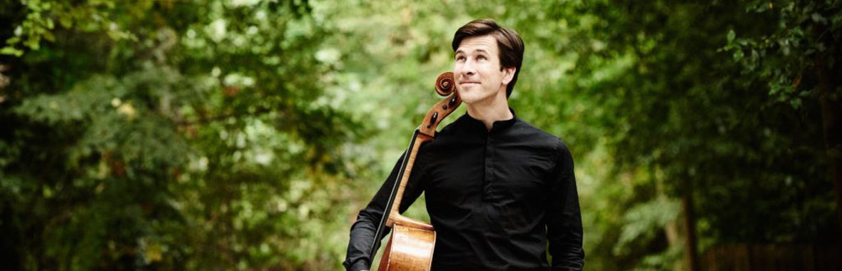 Man standing outside amidst trees and bushes, wearing all black and holding a cello at his side.