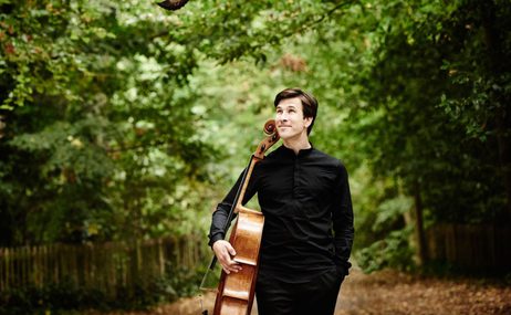 Man standing outside amidst trees and bushes, wearing all black and holding a cello at his side.