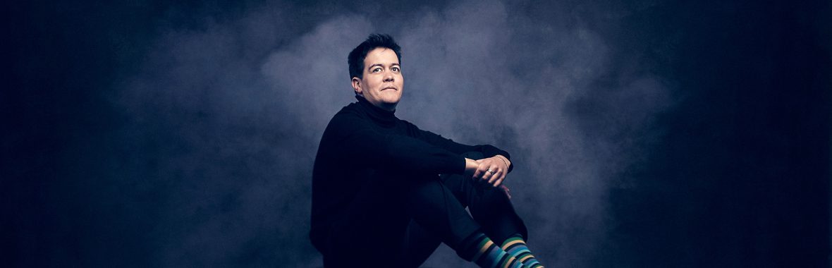 Man with short black hair sitting with his hands folded in front of him.