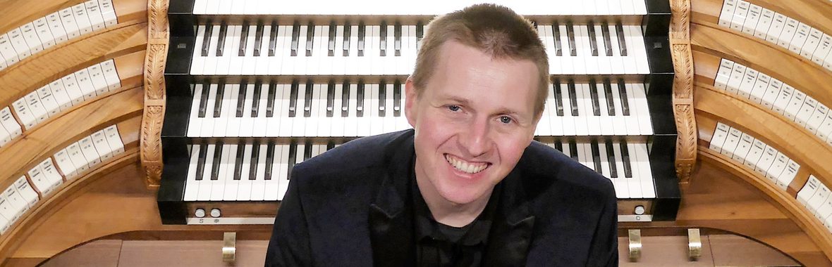 Man with very short hair sitting in front of a church organ and smiling.