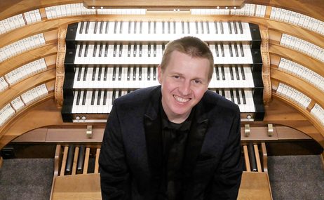Man with very short hair sitting in front of a church organ and smiling.