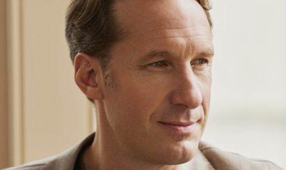 Close-up of a man with short brown hair sitting in front of a window.