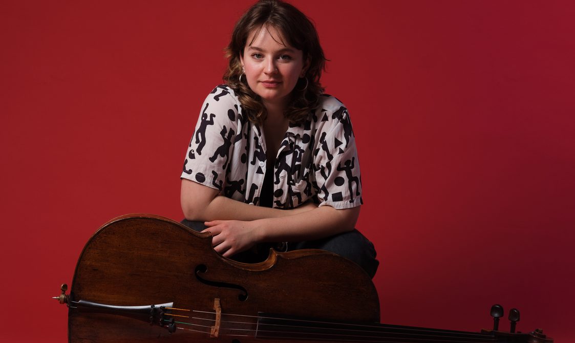 Woman with long brown hair posing with a cello against a red background.
