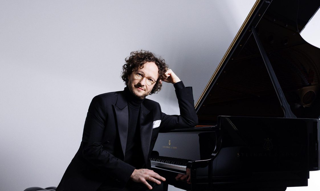 Man with short curly hair and glasses sitting with his arm resting on a grand piano.