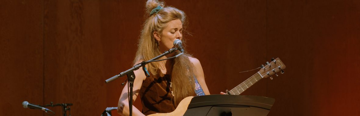 Woman with long blonde hair standing onstage playing a guitar and singing.