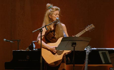 Woman with long blonde hair standing onstage playing a guitar and singing.