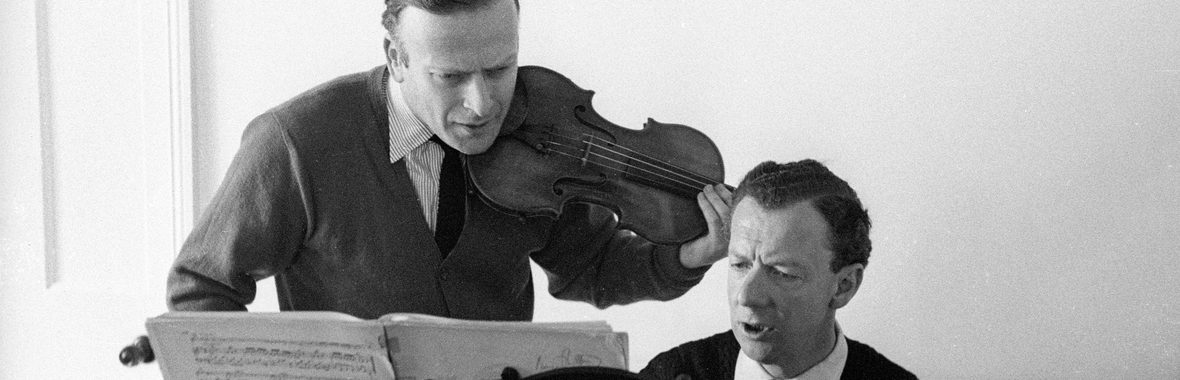 black and white view of two men, one playing the piano and one holding a violin, looking at sheet music together.