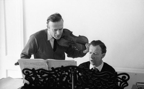 black and white view of two men, one playing the piano and one holding a violin, looking at sheet music together.