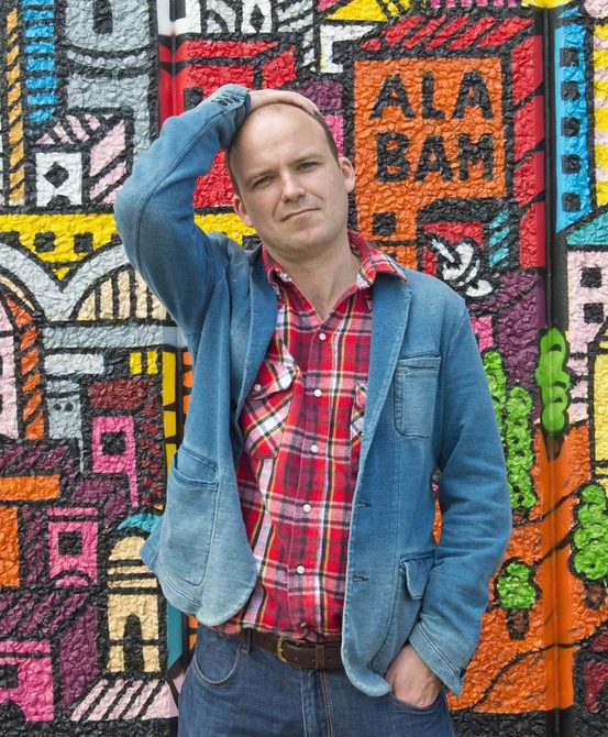 Man with close shaved head wearing a plaid shirt and a denim jacket standing in front of a brightly-patterned wall of art.