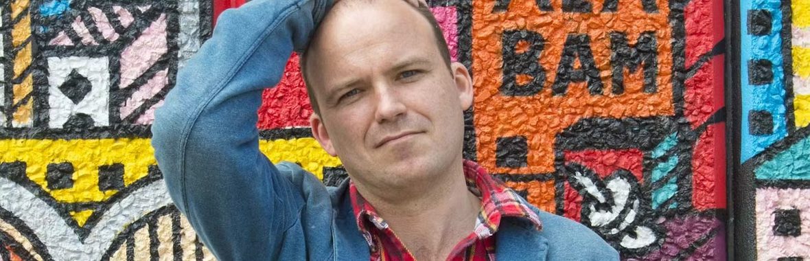 Man with close shaved head wearing a plaid shirt and a denim jacket standing in front of a brightly-patterned wall of art.