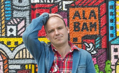 Man with close shaved head wearing a plaid shirt and a denim jacket standing in front of a brightly-patterned wall of art.