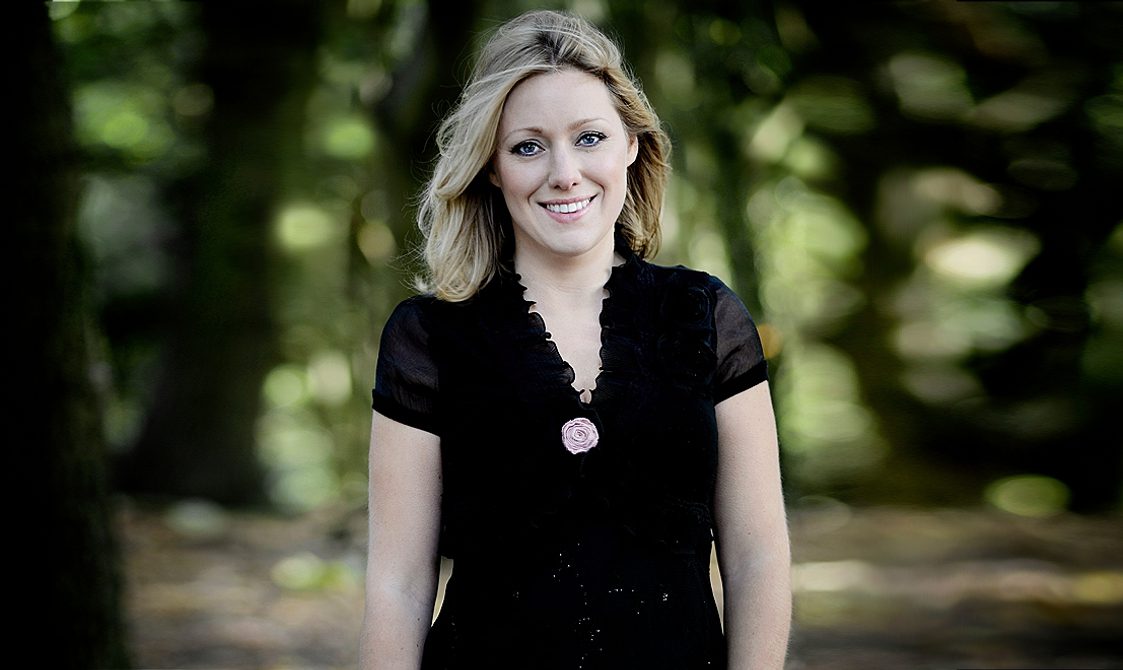 Blonde woman wearing a black dress, standing in front of blurred out trees and smiling.