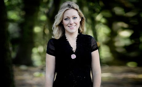 Blonde woman wearing a black dress, standing in front of blurred out trees and smiling.