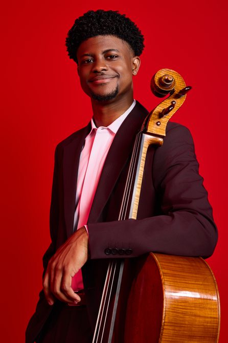 Young man wearing a dark red suit with a pink shirt, holding a cello and smiling.