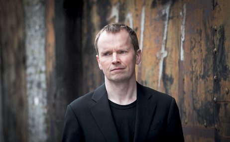 Main with short brown hair, wearing a black jacket with a decorated wall in the background.
