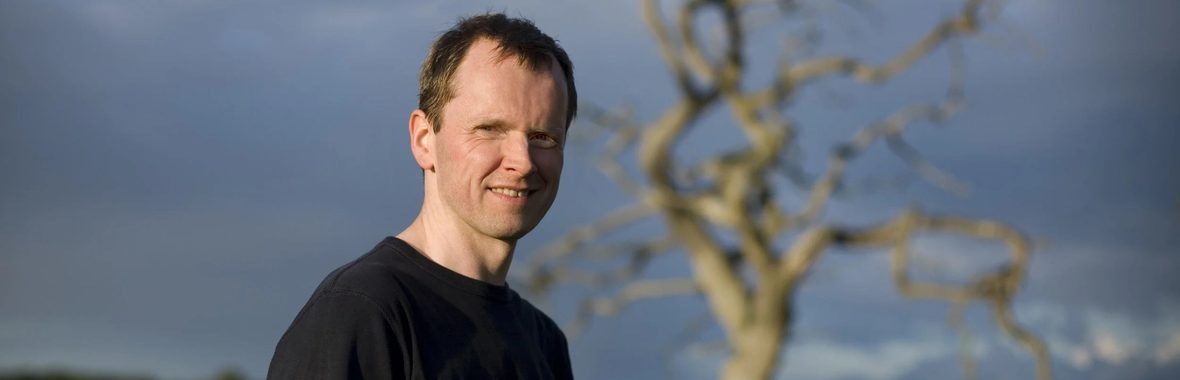 Man with short hair wearing a black t-shirt, a blurred out tree in the background.