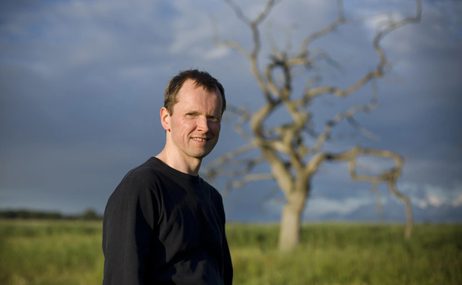 Man with short hair wearing a black t-shirt, a blurred out tree in the background.