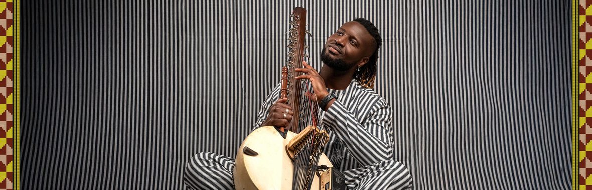 Man wearing striped clothing and sitting with a kora instrument, against a black and white striped background.