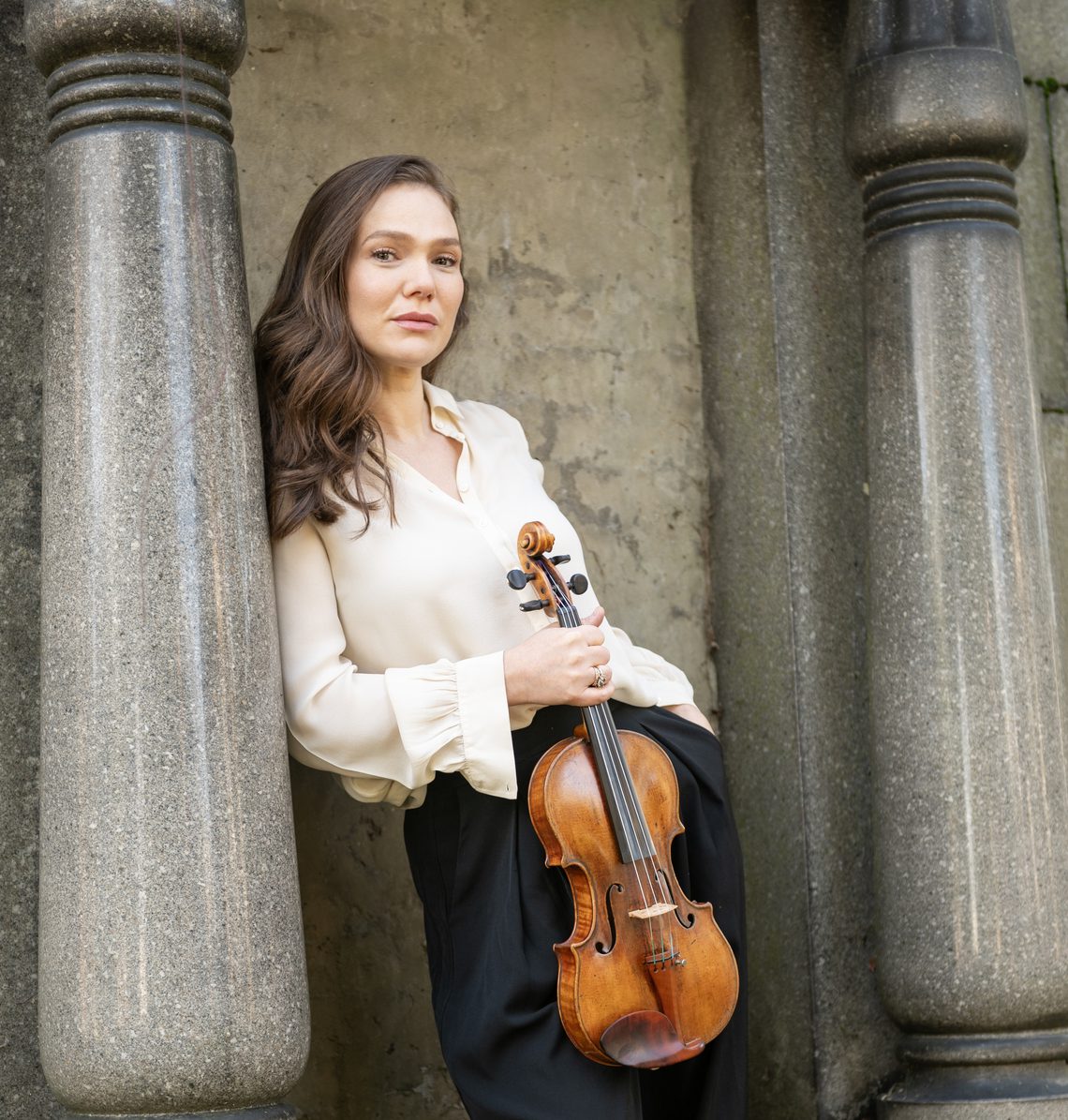 Woman with long brown hair wearing a cream blouse and holding a violin.