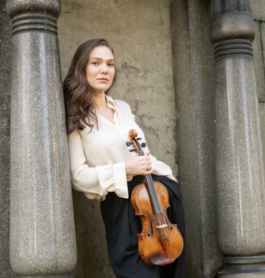 Woman with long brown hair wearing a cream blouse and holding a violin.