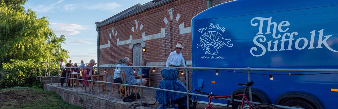 Red brick building with people seated at cafe tables with a horsebox branded The Suffolk in the foreground.
