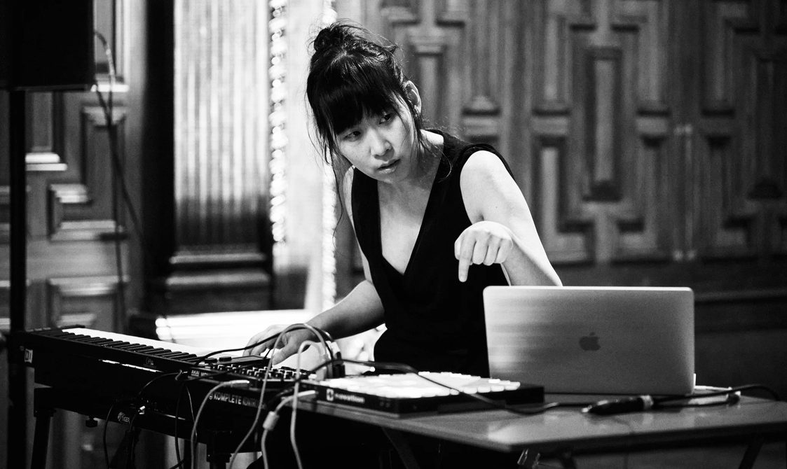 Black and white picture of a woman with black hair tied in a bun working with a keyboard and laptop.