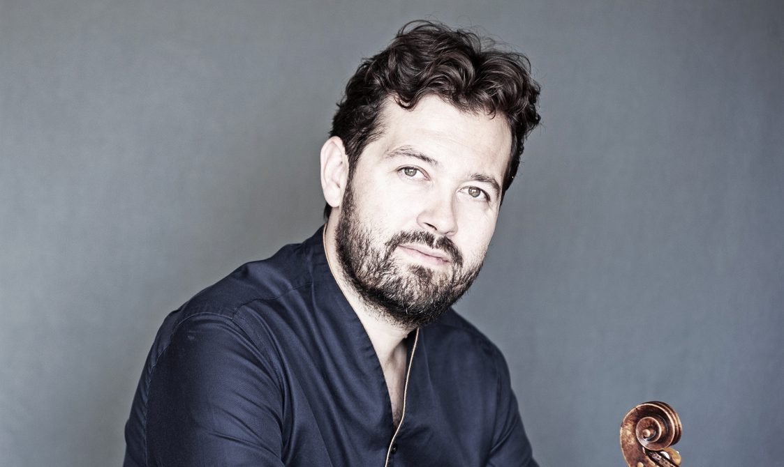 Main with curly black hair and beard wearing a blue jacket and holding a viola.